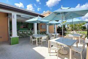 Un patio al aire libre con mesas, sillas, sombrillas y una zona de bar con paredes verdes y una barra.