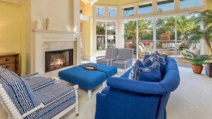 A spacious living room with a fireplace, blue furniture, and large windows offering a view of the outdoors. - Bougainvillea Home (Captiva)
