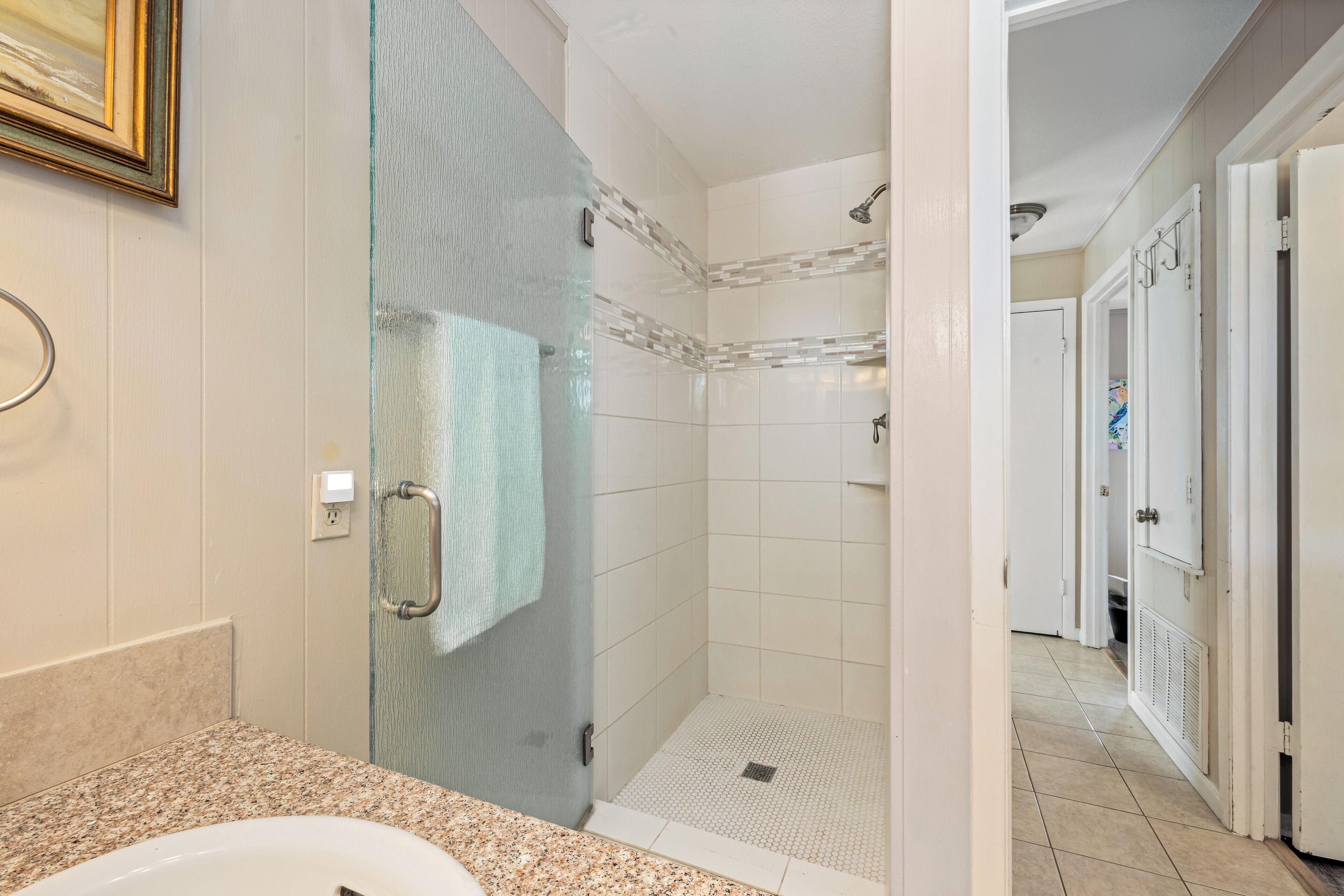 A modern bathroom with a glass-enclosed shower, a granite countertop, and a white tiled floor.