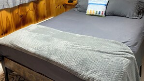 Un dormitorio con pared revestida de madera, una cama con ropa de cama gris, una mesita de noche con lámpara y cortinas grises.