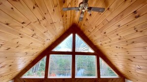 Una habitación con techo inclinado, paneles de madera, un ventilador de techo y cuatro ventanas que ofrecen vista a árboles y un cuerpo de agua.
