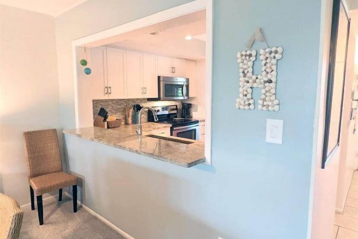A modern kitchen with white cabinets, a microwave, and a coffee maker. There is a chair in the foreground and a decorative letter 'H' on the wall.