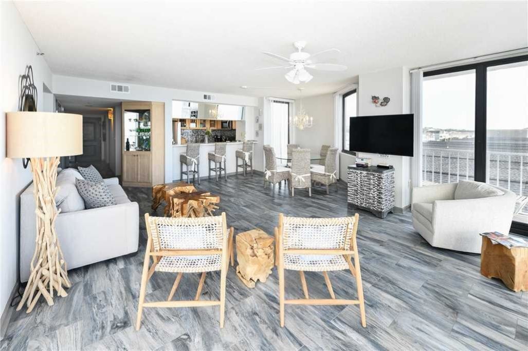 A modern living room with a sofa, two wicker chairs, a coffee table, and a TV. There is a dining area with a table and chairs, and a kitchen in the background.