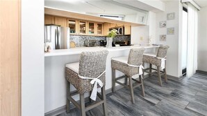 Una cucina moderna con mobili in legno, un frigorifero in acciaio inox e un piano di lavoro bianco. Sono presenti tre sedie in vimini con cuscini bianchi e un asciugamano bianco legato allo schienale.