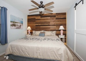 Een slaapkamer met een houten hoofdbord, een bed met een bloemenbedekking, twee bedlampen, een plafondventilator en een schilderij aan de muur.