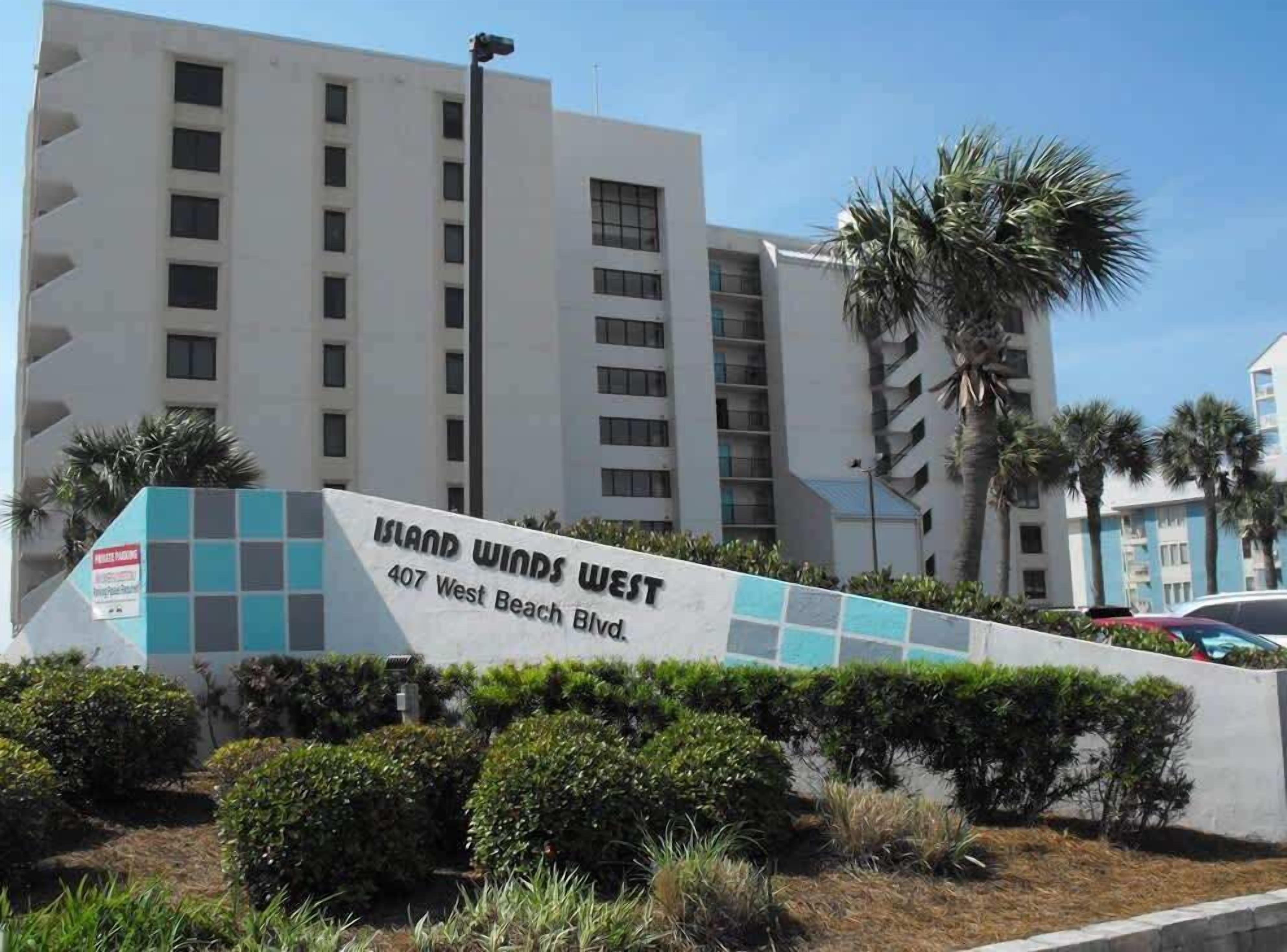 Señal de 'Island Winds West' en el 407 de West Beach Blvd., que muestra un edificio moderno de varios pisos y un cielo despejado.
