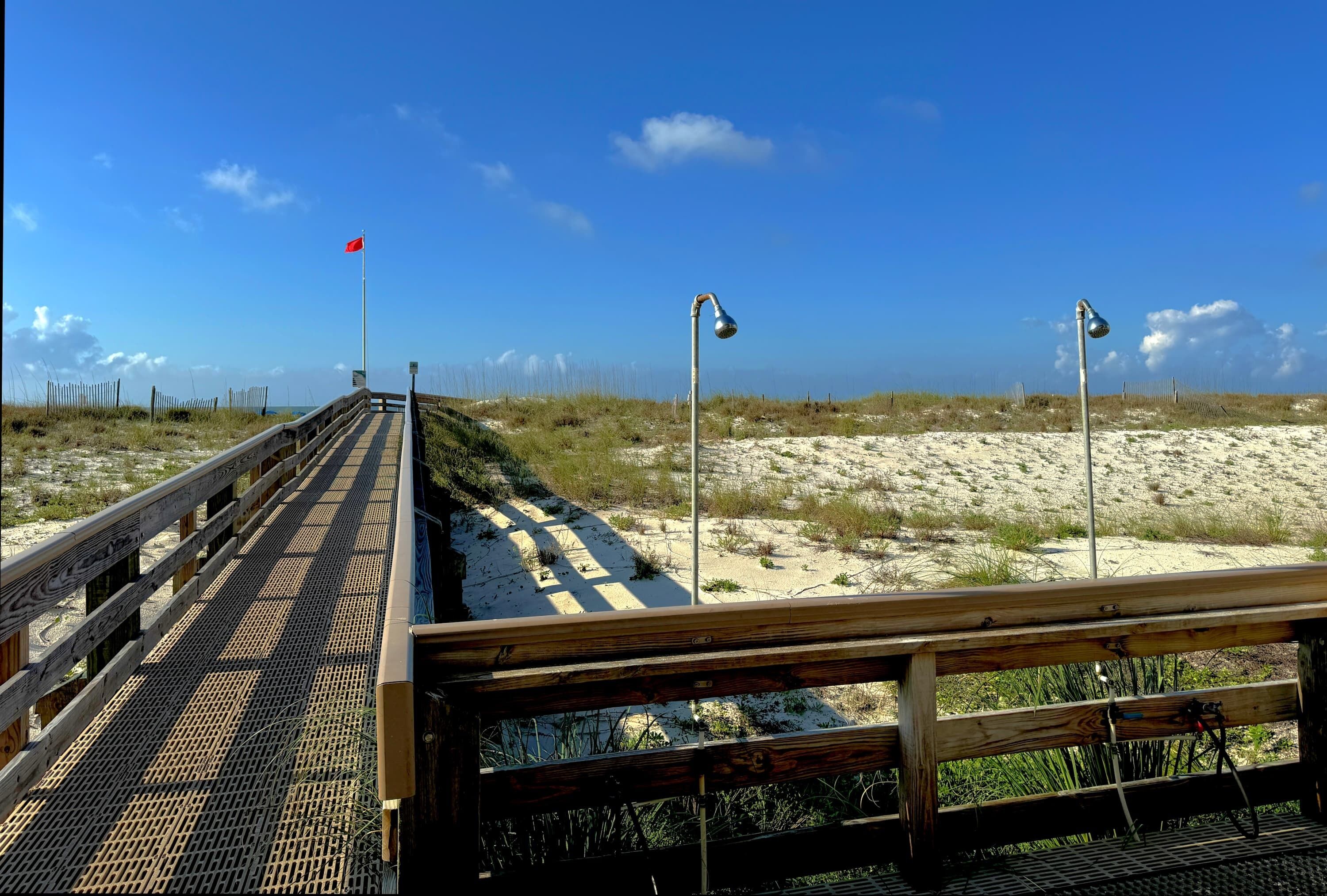 A wooden boardwalk leading to a sandy beach with a flag and street lamps.