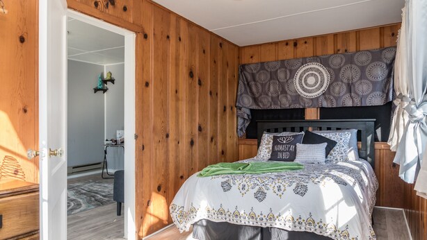 A bedroom with a wooden wall, a bed with patterned bedding, a headboard with a circular design, and a visible door leading to another room. - Zen Den at Rockaway Rentals near Branson (Rockaway Beach)