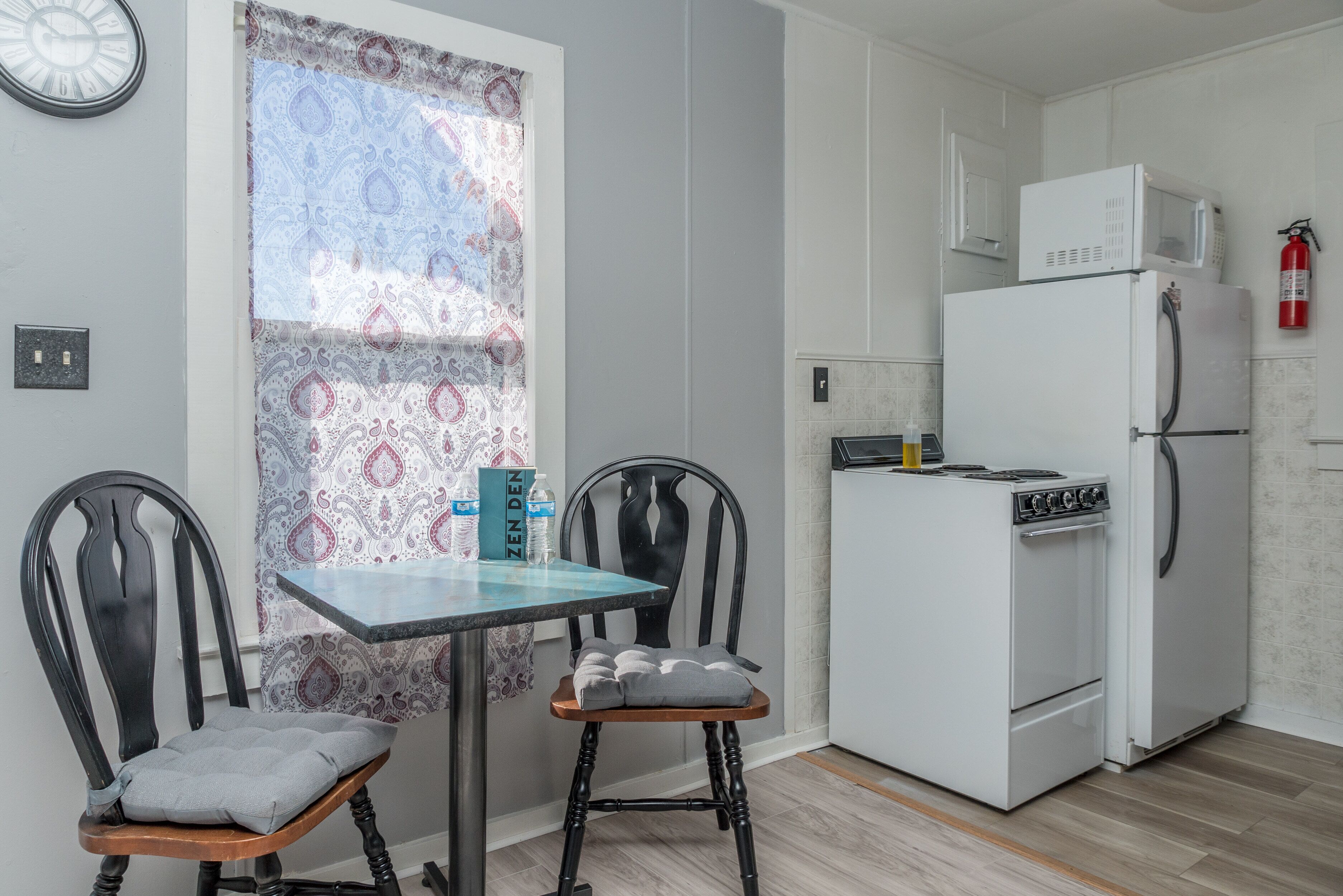 Una cocina compacta con un refrigerador blanco, estufa y microondas. Hay una pequeña mesa con dos sillas y una persiana con un diseño.
