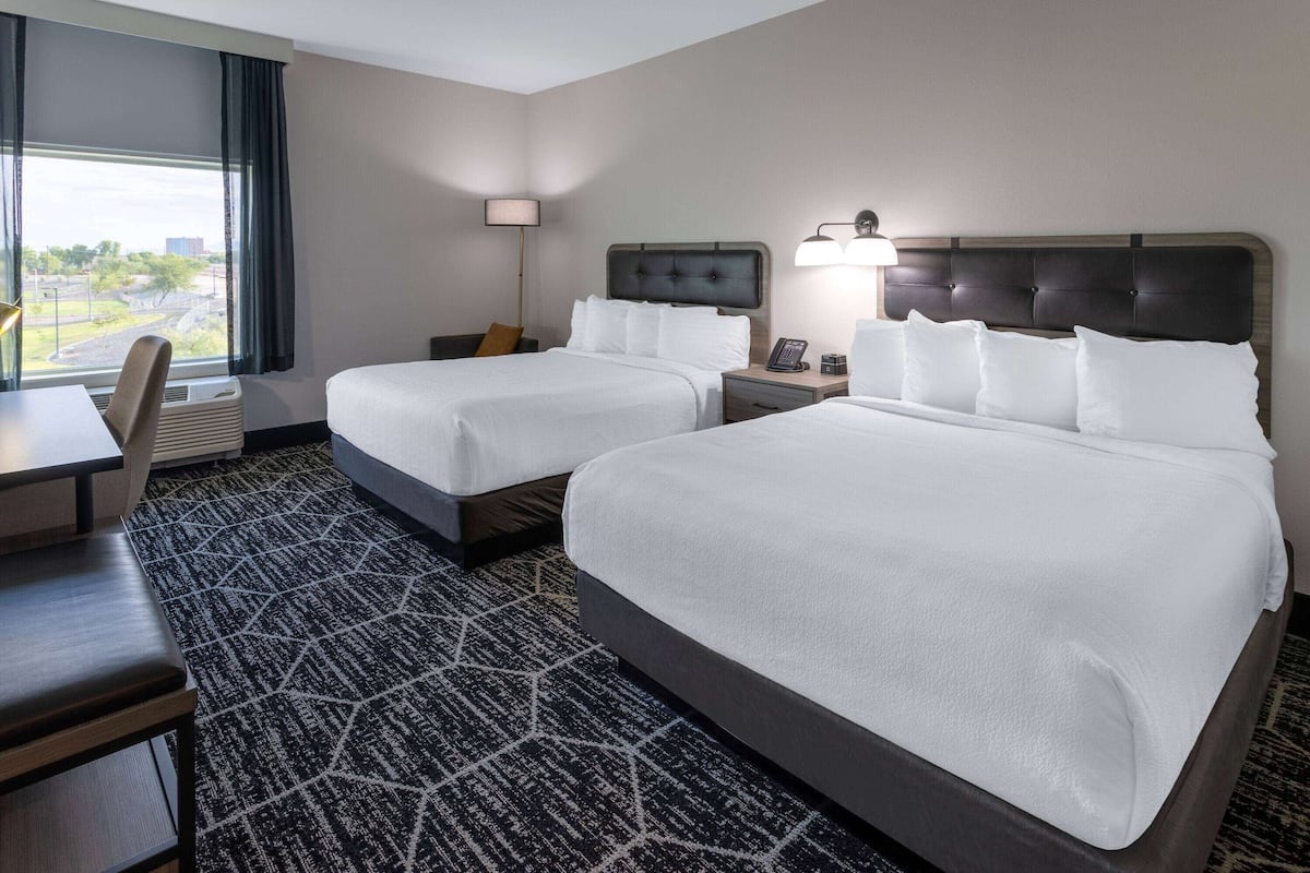 Une chambre d’hôtel avec deux lits, un bureau, une chaise, une lampe et une vue sur l’extérieur.
