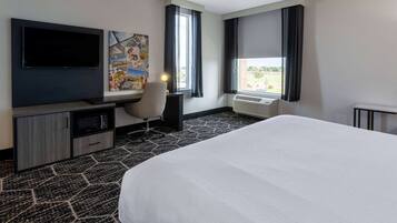 A hotel room with a large bed, a flat-screen TV, a desk with a chair, a wall-mounted air conditioner, and a window with curtains.
