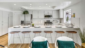 A modern kitchen with white cabinetry, stainless steel appliances, and a central island with bar stools. - Pvt Oasis Pickleball/Basketball/BBQ/Game Room/Pool (Bermuda Dunes)