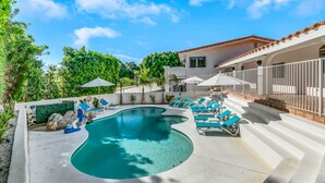 A swimming pool with lounge chairs and umbrellas, surrounded by a white building with a tiled roof and a stone wall. - Pvt Oasis Pickleball/Basketball/BBQ/Game Room/Pool (Bermuda Dunes)