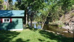 Uma cabana pequena com telhado verde e portaria vermelha, localizada à beira de um rio calmo rodeado por árvores.