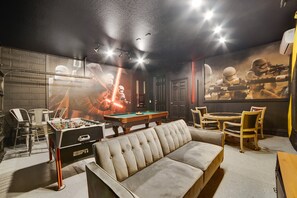 A room with a foosball table, a pool table, a sofa, and a Star Wars-themed wall mural.