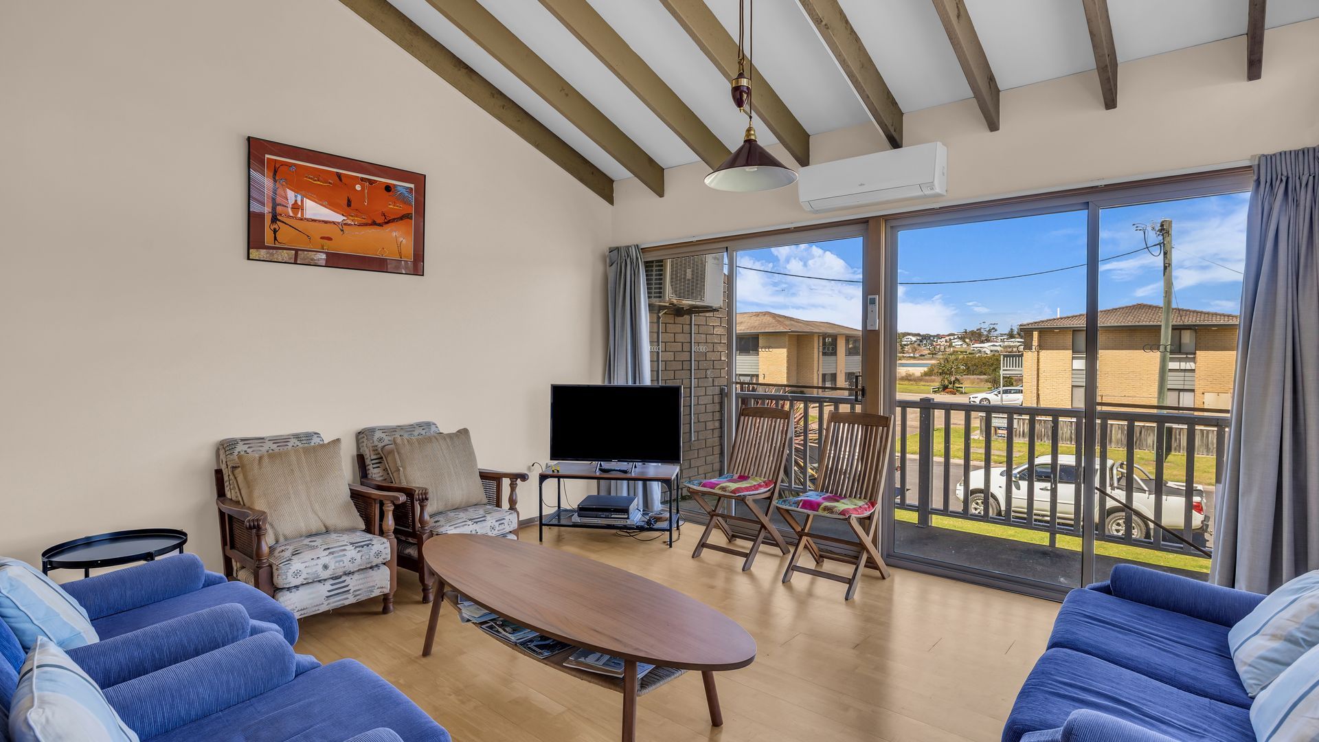 A living room with a TV, sofas, a table, and a balcony with a view of houses.