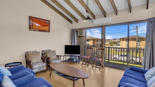 A living room with a TV, sofas, a table, and a balcony with a view of houses.