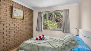 A bedroom with a brick wall, a window with curtains, a bed with a green blanket, and a painting on the wall.