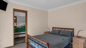 A bedroom with a wooden bed frame, a nightstand, a drawer unit, and a bathroom visible through an open door. - Outlook 1 - Unit 1 Narira St 9 (BERMAGUI)