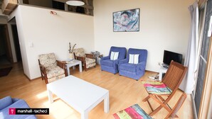 A living room with a white table, two blue armchairs, a wooden chair, a small white table, and a colorful rug. - Outlook 1 - Unit 1 Narira St 9 (BERMAGUI)