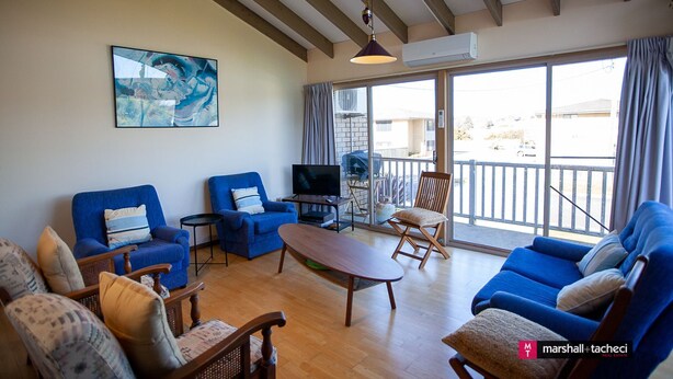 A spacious living room with a large TV, blue sofas, and a balcony with a view. - Outlook 1 - Unit 1 Narira St 9 (BERMAGUI)