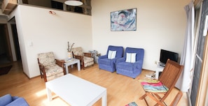 Un salon avec une table blanche, deux fauteuils bleus, une chaise en bois, une petite table blanche et un tapis coloré.