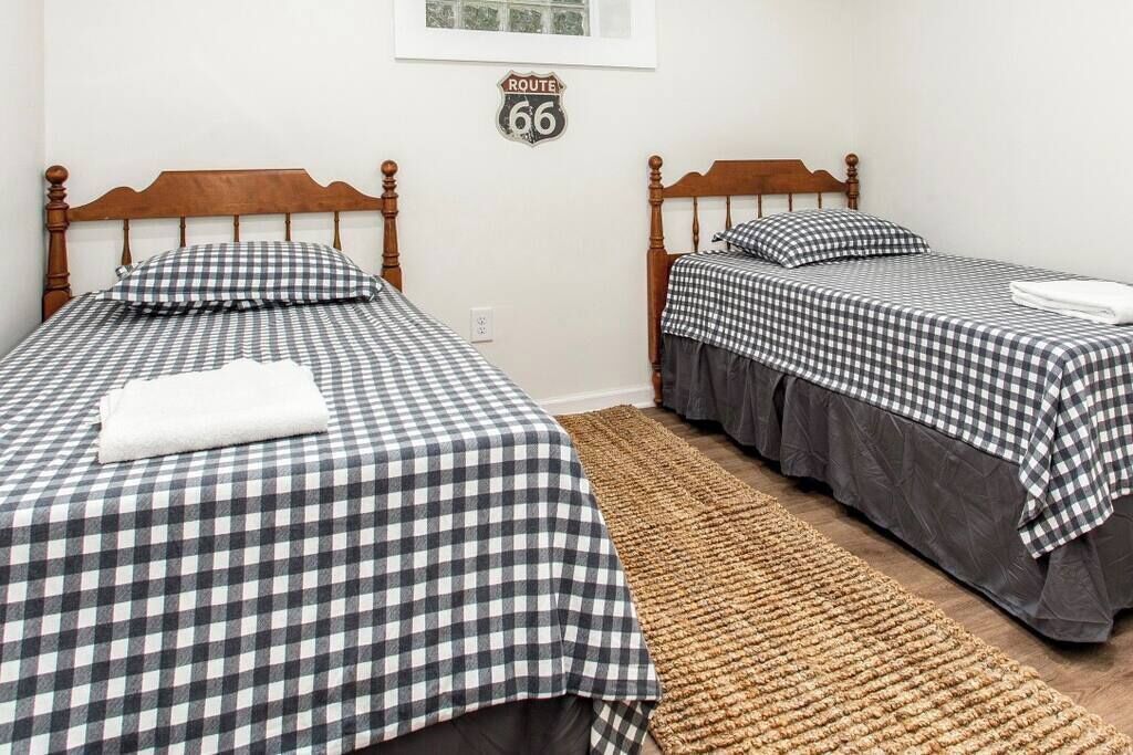 Una habitación con dos camas de madera, cada una con colcha a cuadros y almohadas a juego. Un tapiz tejido cubre el suelo, y un cartel de 'Route 66' está en la pared.