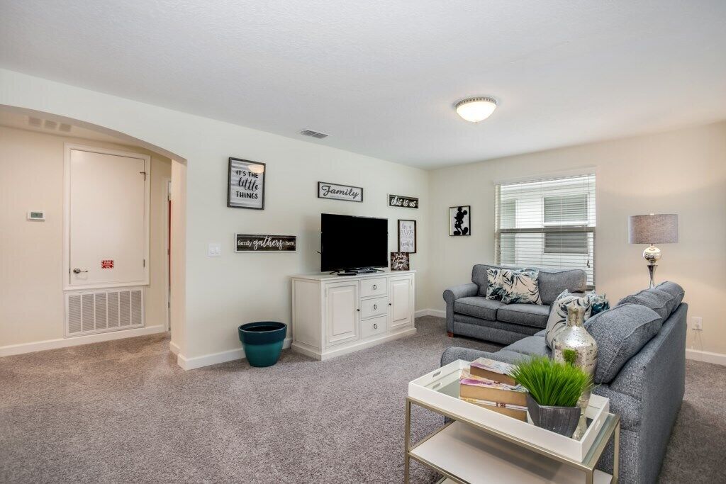 Una sala de estar con un sofá gris, un mueble blanco para equipo multimedia y un televisor. Se observan decoraciones en las paredes y una ventana con persianas.