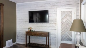 A room with a wooden wall, a mounted TV, a wooden console table with drawers, a door with a herringbone pattern, and a lamp on a side table.