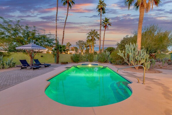 A swimming pool with a curved edge, surrounded by palm trees and cacti, with lounge chairs and a shaded area in the background. - Olive Tree Retreat | Heated Pool, Patio & Privacy (Scottsdale)