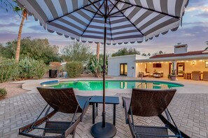 A poolside area with a covered seating area, a pool, and a building in the background. - Olive Tree Retreat | Heated Pool, Patio & Privacy (Scottsdale)