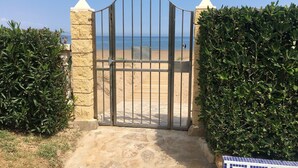 Une entrée fermée par un portail en métal et bordée d’un mur de pierre, qui mène à une plage.