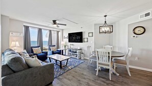 A modern living room with a sofa, coffee table, and dining area. There is a large window with a view of the ocean.