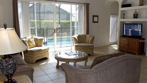 Ein Wohnzimmer mit einer Couch, zwei Sesseln, einem Couchtisch, einem Fernseher und einer Glasschiebetür, die zu einem Pool führt.