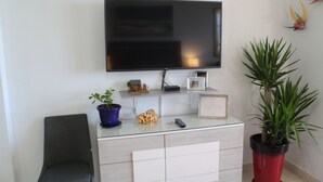 A living room with a flat-screen TV mounted on the wall, a grey sofa, a glass coffee table with a potted plant, a red potted plant, and a framed certificate. - San Juan Sunrise - Apex Living in the Capital of Puerto Rico (San Juan)
