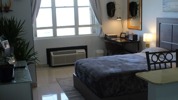 A modern hotel room with a large bed, a desk with a chair, a desk lamp, and a large window with curtains. - San Juan Sunrise - Apex Living in the Capital of Puerto Rico (San Juan)