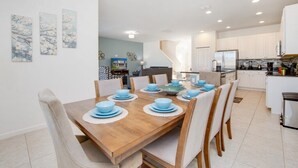 Un moderno comedor con una mesa de madera preparada para seis personas, rodeada de sillas. Se ve una cocina al fondo con armarios blancos y un refrigerador de acero inoxidable.