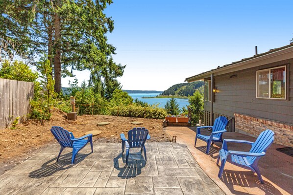 A patio with four blue Adirondack chairs, a stone fireplace, and a view of a lake and hills.