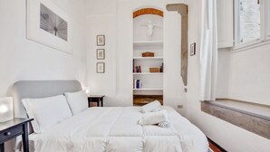 Habitación de hotel con cama, mesita de noche, estante en la pared con libros y una estatua, y ventana con cortinas.
