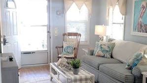 A living room with a sofa, armchair, and a small table with a plant. There is a door leading outside and a window with curtains. - Ocean Lakes Pelican Pointe, Beautiful Ocean & Lake Front cozy cottage. (Myrtle Beach)