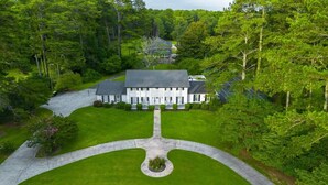 A large, white, two-story house with a symmetrical facade, surrounded by lush greenery and a central driveway leading to the entrance.