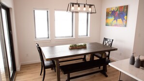 A dining area with a wooden table, benches, and a chandelier. There are windows with blinds, a colorful painting on the wall, and a small plant on the table.