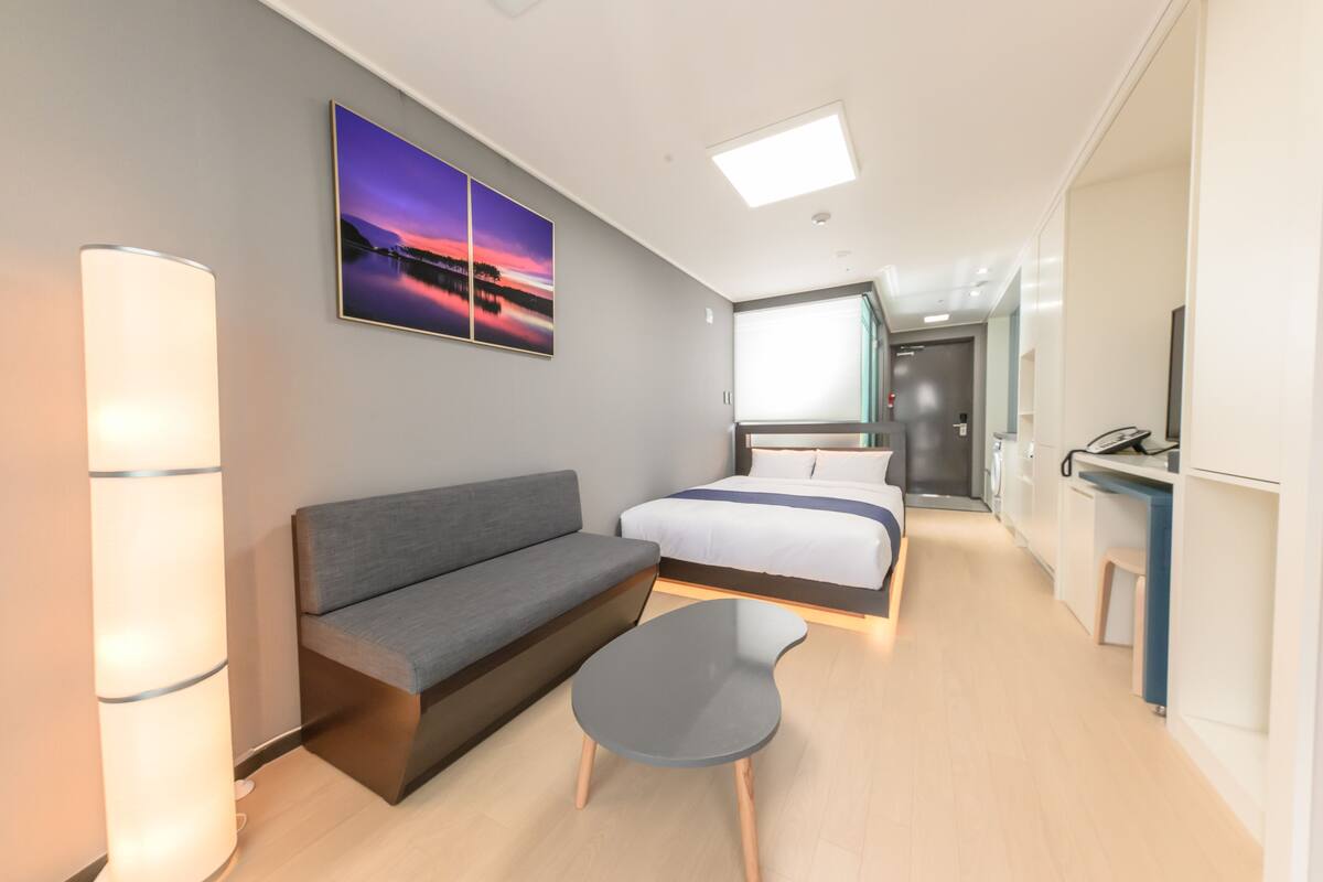 Quarto de hotel moderno com sofá, mesa pequena, cama, escrivaninha e uma janela grande.