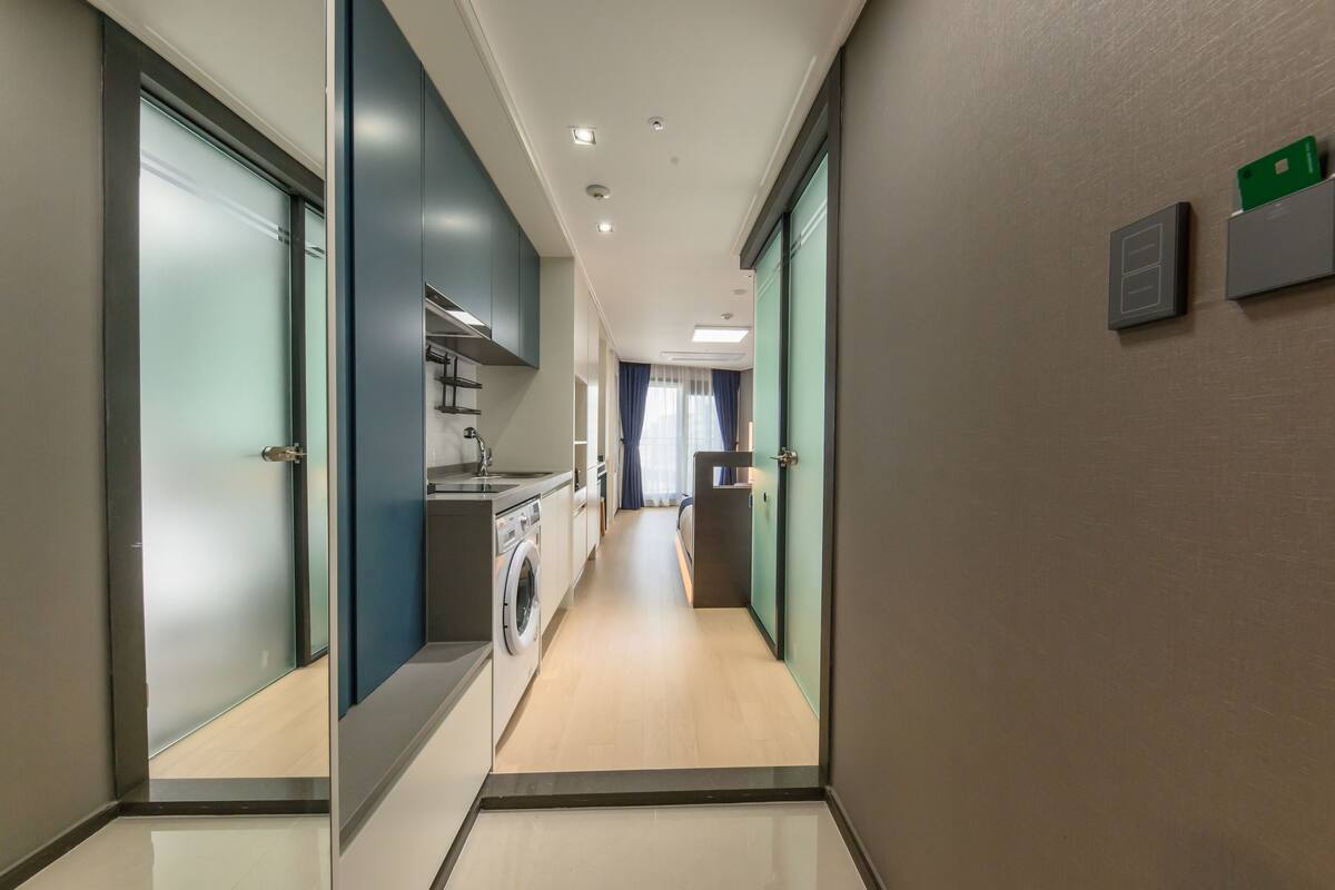 Corredor moderno de hotel com cozinha, incluindo máquina de lavar, e vista para um quarto com cama e cortinas.