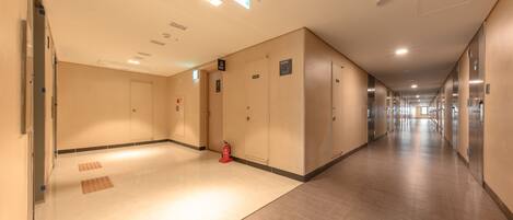 Un largo pasillo bien iluminado, con paredes color beige y piso de madera, que cuenta con varias puertas y un extintor.