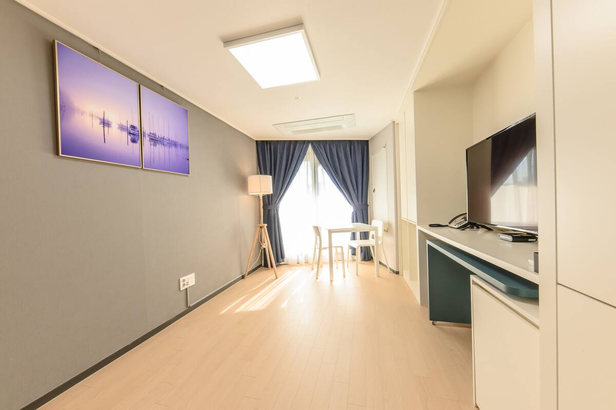 Quarto de hotel moderno com uma televisão de tela plana grande, uma escrivaninha com cadeira, uma luminária e um quadro na parede.