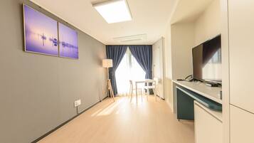 A modern hotel room with a large flat-screen TV, a desk with a chair, a lamp, and a painting on the wall.
