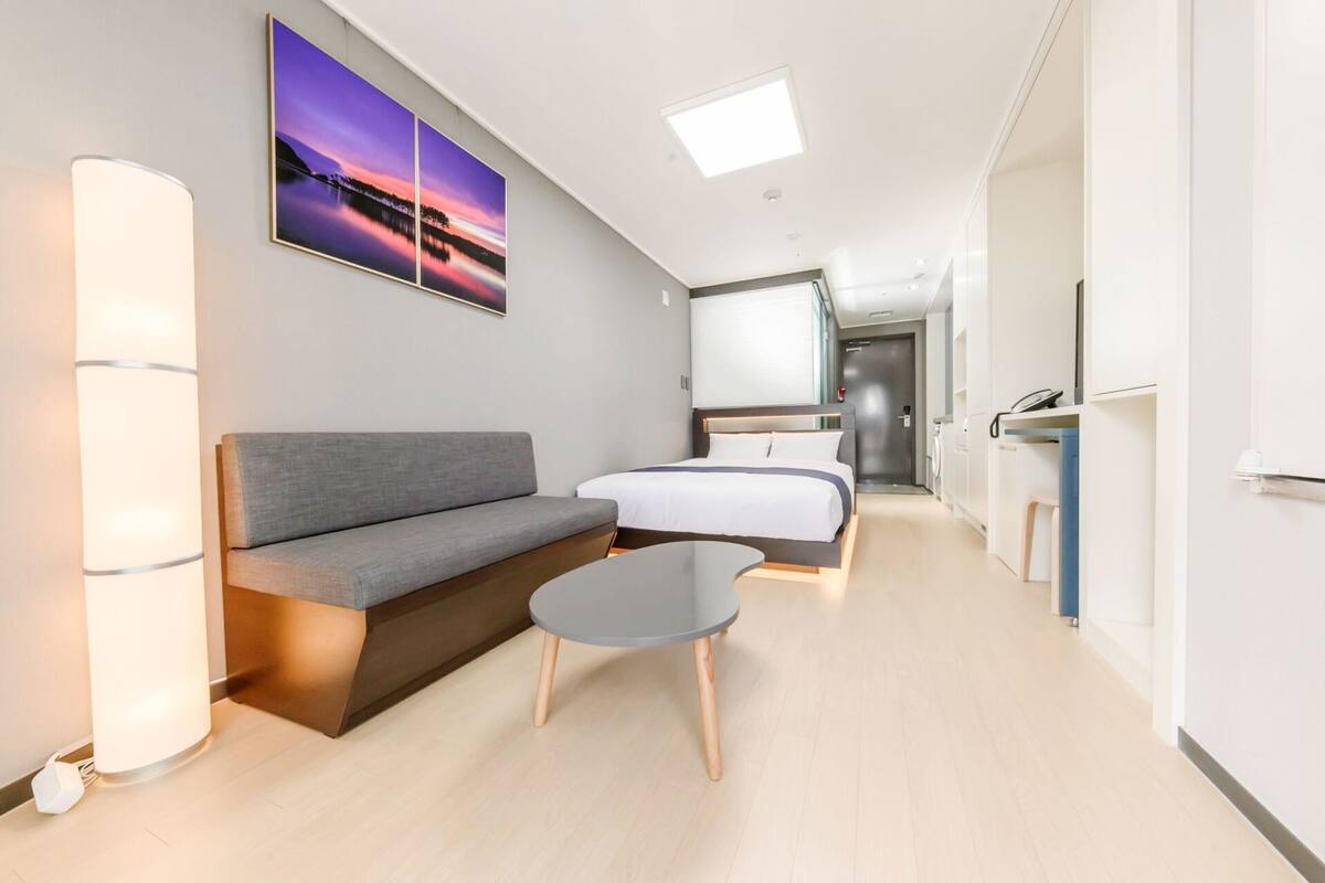 Quarto de hotel com sofá, cama, mesa pequena, mesa de cabeceira, escrivaninha, cadeira, espelho e um quadro na parede.