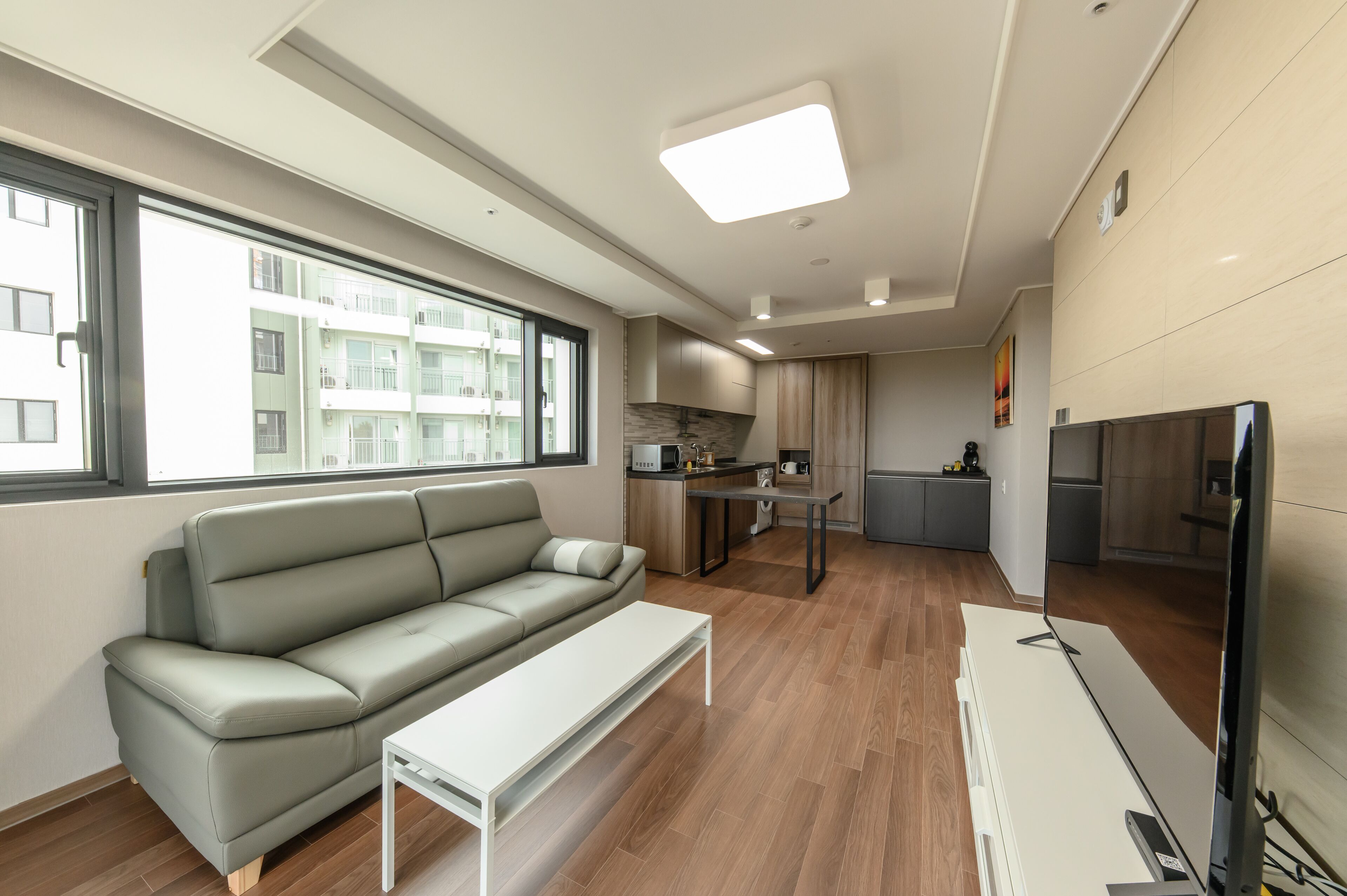 Un apartamento moderno con un sofá gris, una pequeña mesa blanca, una zona de cocina y un televisor grande.