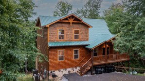 A two-story log cabin with a green metal roof, a wooden deck, and a stone foundation. - New! “WaterTastic” Private Cabin heated Indoor Pool, Theater, fantastic view  (Gatlinburg)